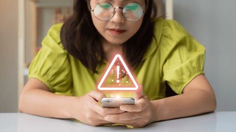 A woman in a lime green top focuses on her smartphone, which displays a caution symbol, highlighting the importance of online security awareness.