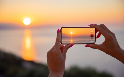 Hands holding a smartphone capturing the sunset over the sea, showcasing landscape photography.