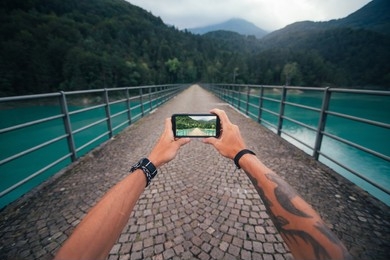 A person standing on a cobblestone bridge over a turquoise river captures the scenic view with a smartphone camera.