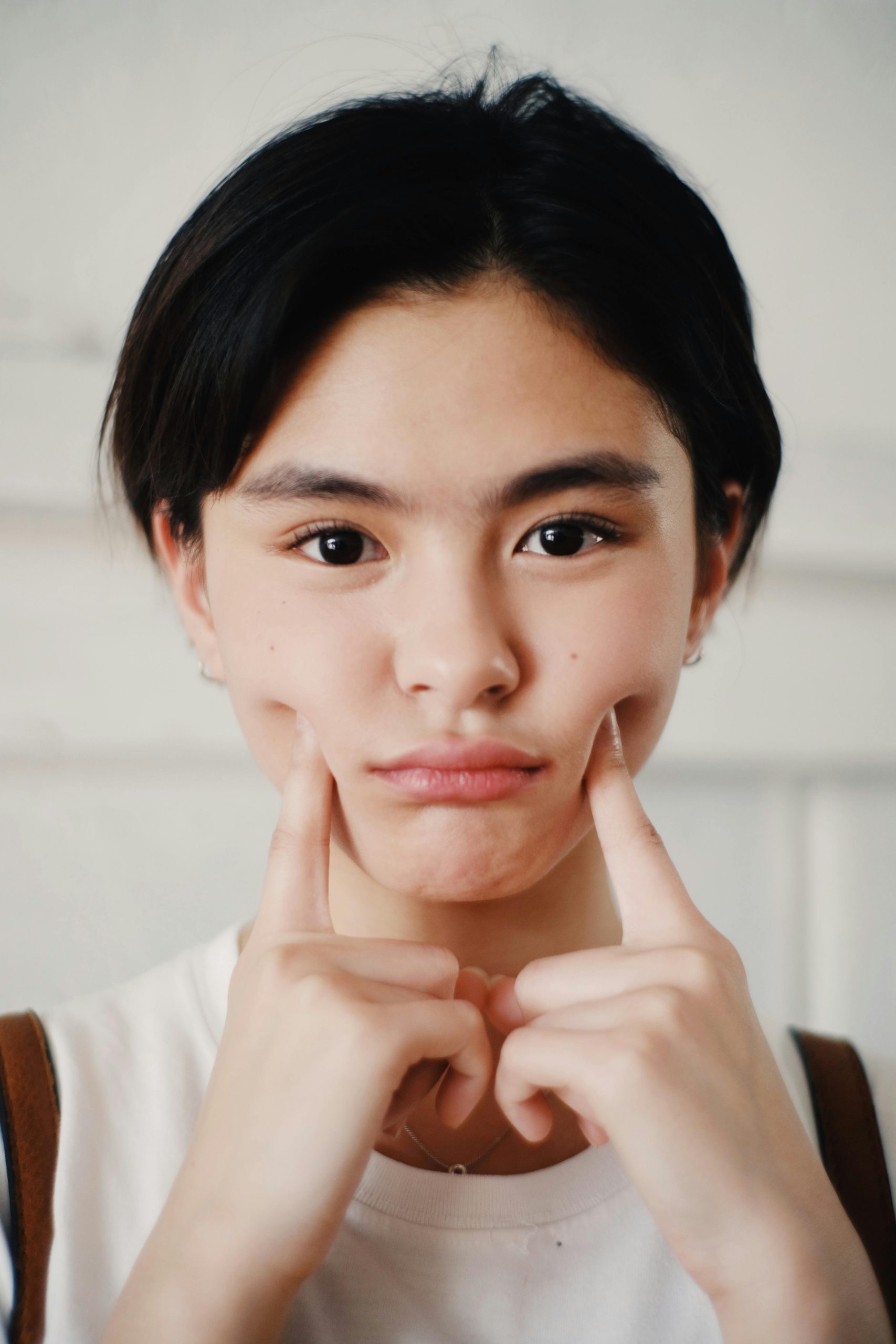 A young woman with black hair and a neutral expression, playfully forcing a smile by pushing up the corners of her mouth with her fingers, against a soft-focused white background.