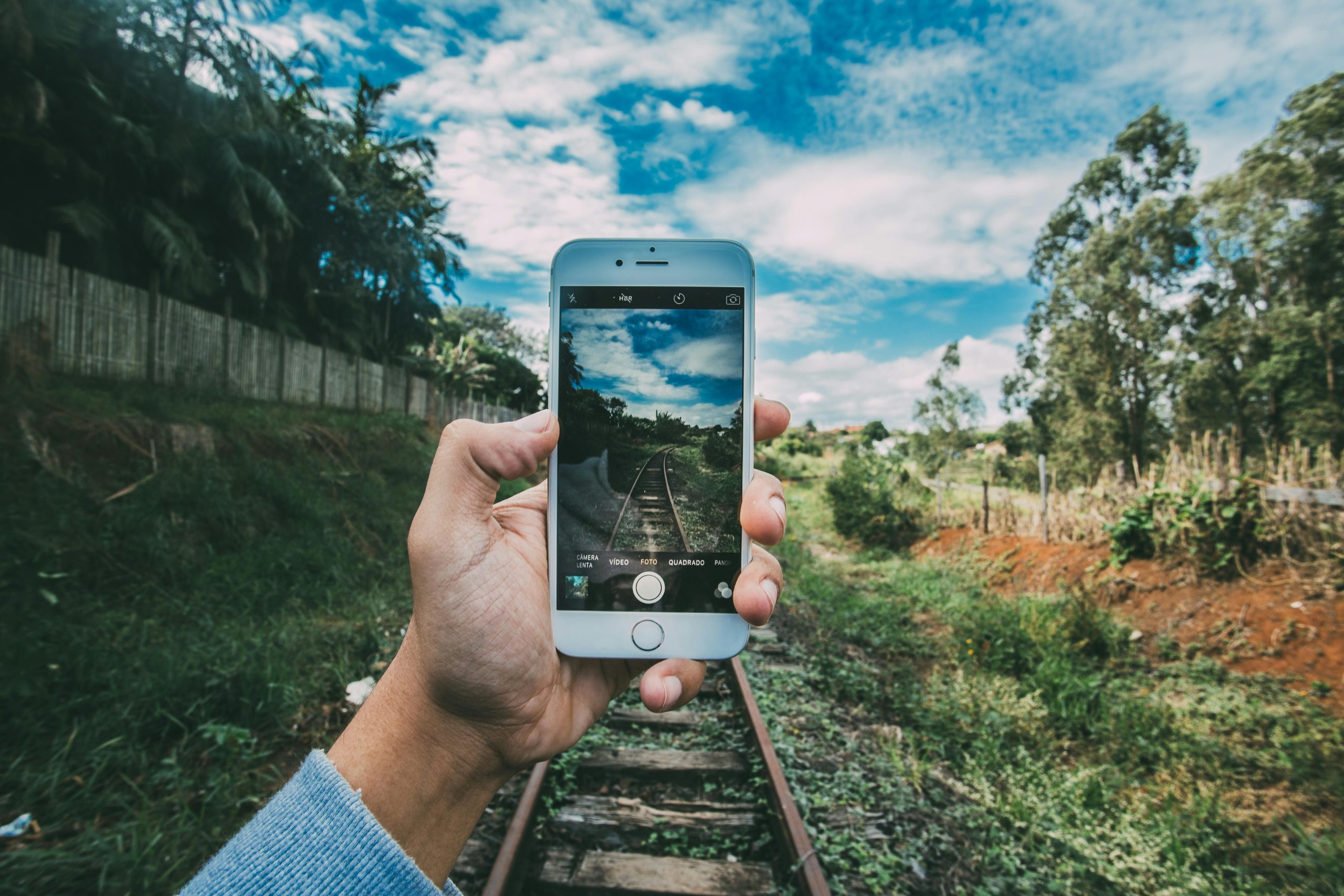 A hand taking a picture, focusing on a railway track set within a lush, green landscape.