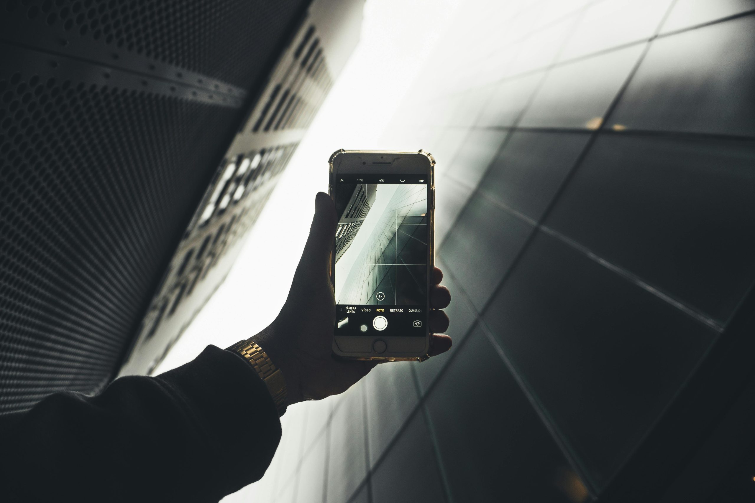 A hand holds a smartphone, photographing the architectural symmetry of a metallic building façade, bathed in soft backlighting.