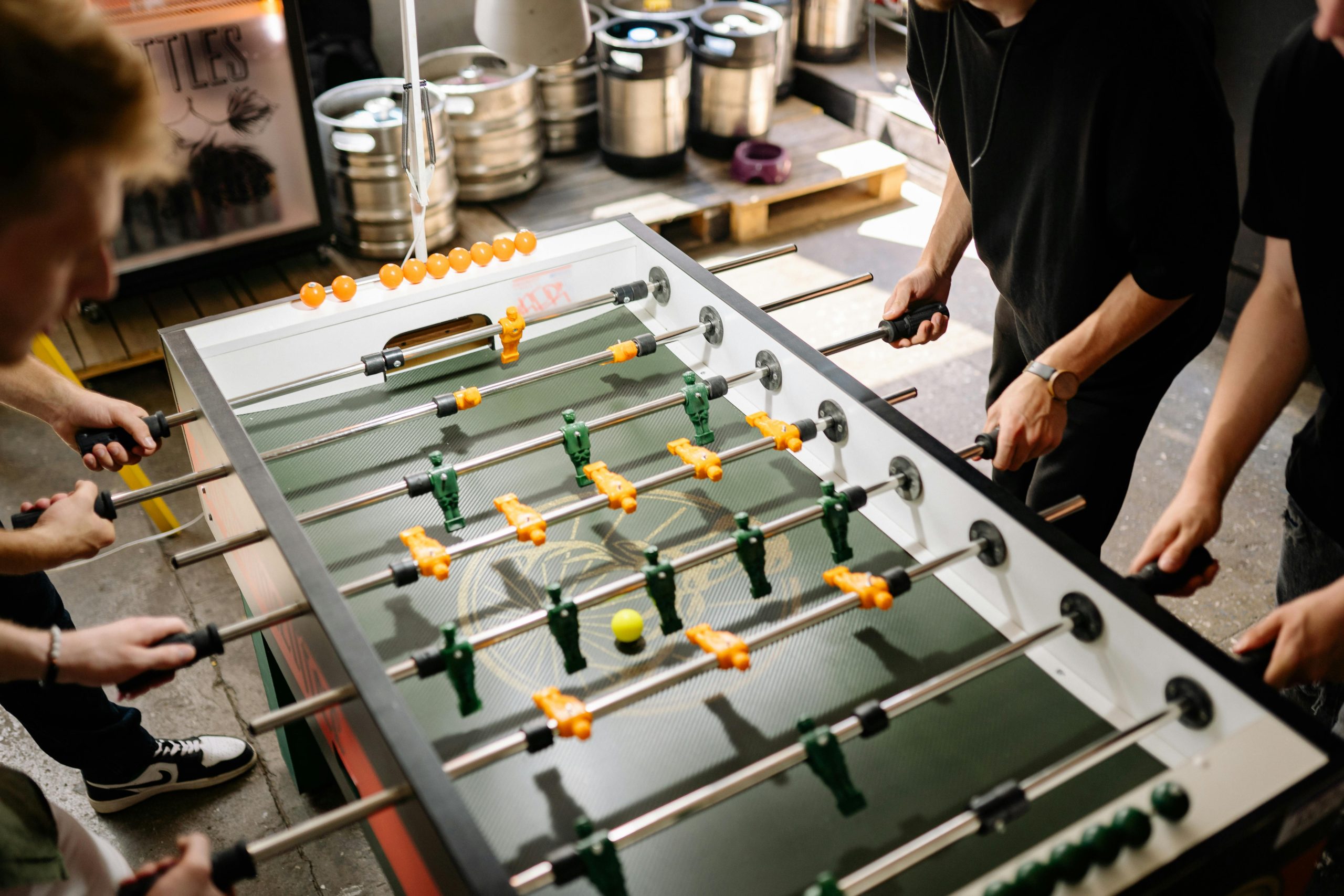 An aerial view of three people engaging in a competitive game of foosball on a standard table, with a bright yellow ball in play.